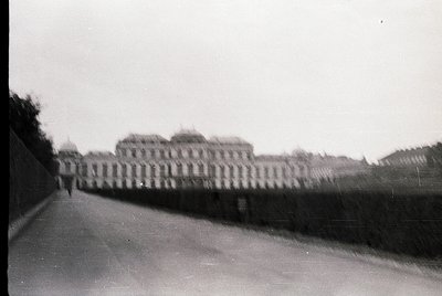 Blurred black-and-white shot of a grand, symmetrical palace façade with arched windows and a high wall, likely European. Over...