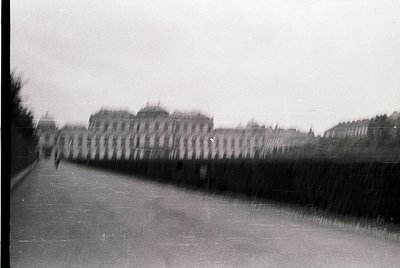 Blurred monochrome street scene with indistinct figures walking alongside a high, dense hedge. Distant buildings suggest urba...