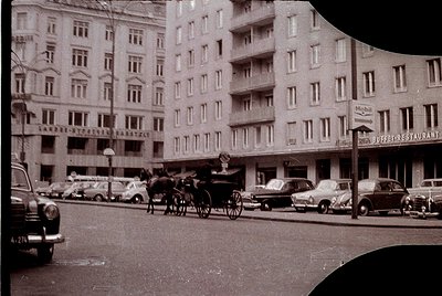 Vintage street scene featuring early 20th-century European urban architecture. Horse-drawn carriage and vintage cars share th...