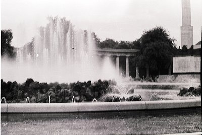 Classic black-and-white shot of a grand **fountain** in an urban plaza, framed by **classical columns** and **monumental tree...