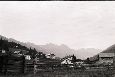 Vintage black-and-white alpine village scene with wooden chalets and rustic fencing. A man in mid-20th-century attire poses n...