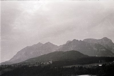Vintage black-and-white mountain landscape with mist-covered peaks and sparse vegetation. Foreground shows a valley with scat...