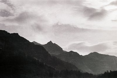 Black-and-white aerial view of rugged alpine peaks with dense forest cover, likely captured mid-20th century. Dramatic cloud ...