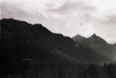 Vintage black-and-white mountain landscape with dense forest cover and misty peaks, evoking a moody, atmospheric feel. Likely...