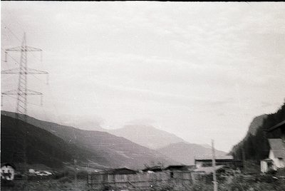 Black-and-white rural landscape featuring a high-voltage pylon on left, spanning across a misty valley. Low-lying buildings a...