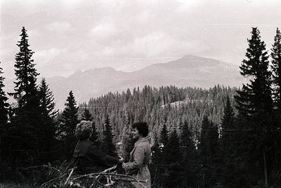 Two individuals in mid-20th-century attire stand on a forested alpine ridge, framed by dense coniferous trees. The distant mo...