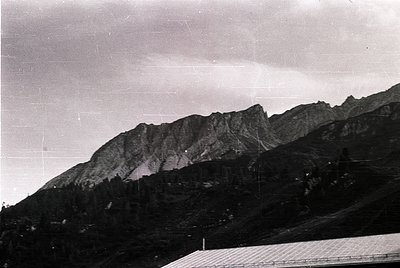 Mid-century black-and-white alpine landscape with jagged peaks and sparse vegetation. Foreground shows a sloped, partially bu...