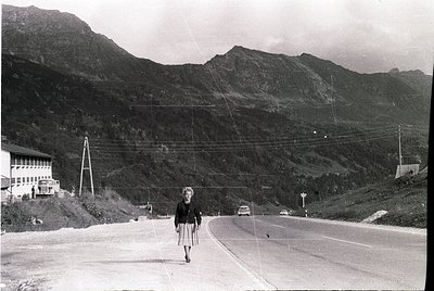 Mid-20th century alpine roadside scene: woman in 1950s-style dress crossing a winding mountain road flanked by utility poles....