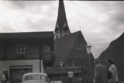 Black-and-white alpine village scene featuring a timber-framed chalet and stone church with a steep spire. Classic vintage ca...