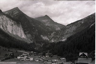 Mid-20th century alpine village nestled between steep, forested slopes and jagged peaks. Classic wooden chalets with pitched ...