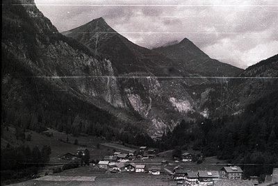 Aerial black-and-white view of a mountainous Alpine village nestled in a valley, likely mid-20th century. Clustered wooden ch...