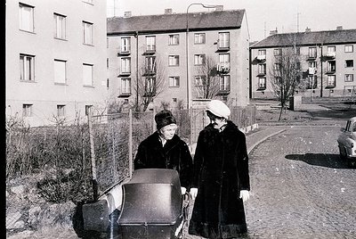 Two women in winter attire push a baby stroller along a cobblestone path beside Soviet-era apartment blocks, 1960s–1970s. Fun...