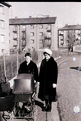 Two women in 1950s-60s winter attire stand beside a vintage pram on a cobblestone street. Brick apartment buildings with balc...