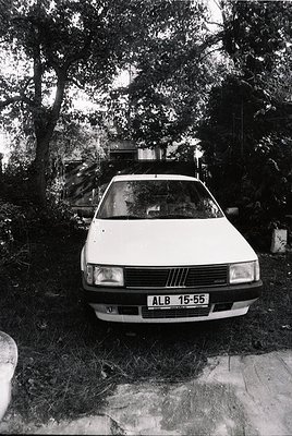 Vintage Fiat 128 sedan (ALB 15-55 license) parked in a wooded driveway, surrounded by dense foliage. Mid-20th century Europea...