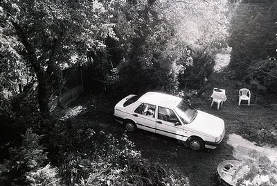 Aerial shot of a 1980s-era sedan parked on a sloped driveway surrounded by dense foliage. Two white plastic chairs and a tabl...
