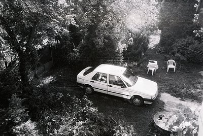 Aerial view of a mid-20th-century sedan parked in a shaded backyard, surrounded by dense foliage. Two white plastic chairs an...