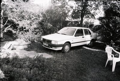 Vintage sedan parked on overgrown grass beside a concrete slab and plastic chair. Likely a 1980s European model, suggested by...