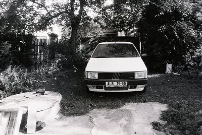 1970s-era Fiat 128 parked on concrete driveway, identifiable by "ALB 15-55" license plate. Surrounded by overgrown foliage an...