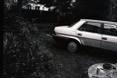 Vintage sedan parked on overgrown grass beside a rusted manhole cover, framed by dense foliage and a distant house. Classic a...
