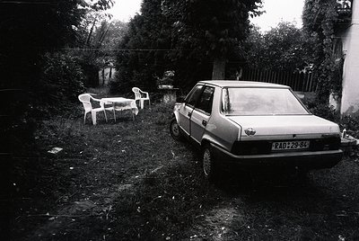 Vintage black-and-white shot of a neglected suburban yard: a faded white sedan (RAD-29-84) parked beside overgrown grass, wit...