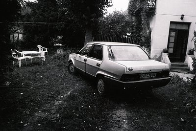 Vintage sedan parked in overgrown backyard with faded white plastic chair and table. Rustic residential setting, likely Easte...