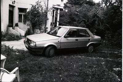 Vintage sedan parked on overgrown grass beside a residential building, likely Eastern Bloc-era design. Noteworthy details: re...