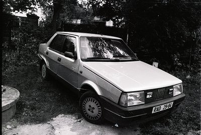 Classic hatchback sedan parked in a wooded area, likely a Fiat 127 based on grille design. Black-and-white photo captures 197...