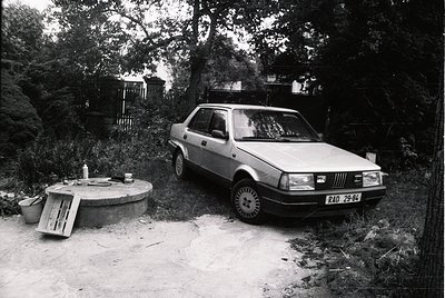1970s-era Fiat 128 parked beside a rustic concrete picnic table and metal bucket. Dense forest and rocky terrain suggest a ru...