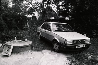 A 1970s-era hatchback parked beside a rustic stone well on a gravel path, surrounded by dense forest. The car’s license plate...