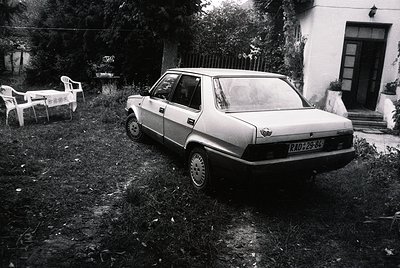 Vintage sedan (RAD-29-86) parked on overgrown grass beside a residential building, likely Eastern Bloc-era. White plastic cha...
