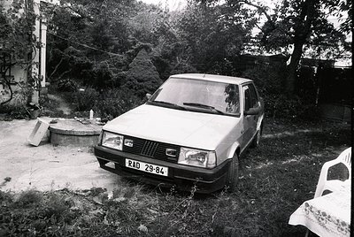 1980s-era Fiat sedan (RAD 29-84) parked in overgrown backyard, surrounded by concrete slab and rusted metal barrel. Residenti...