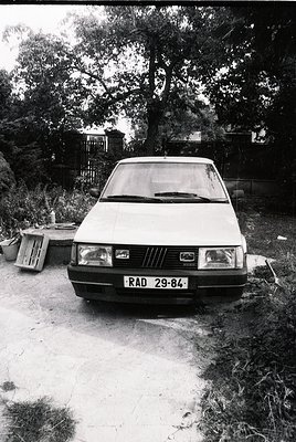 Vintage sedan (likely Fiat 127) parked on gravel, license plate "RAD 29-84" suggests Eastern Bloc (1980s). Rustic backyard wi...
