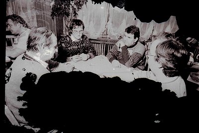 Vintage black-and-white photo of a group seated around a round table in a dimly lit indoor setting, likely a 1960s-70s gather...