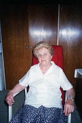 Portrait of an elderly woman seated in a red upholstered armchair, holding armrests for support. She wears a white blouse and...