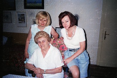 Three generations pose indoors, likely 1980s–1990s. Elderly woman seated in vintage armchair, flanked by a middle-aged woman ...