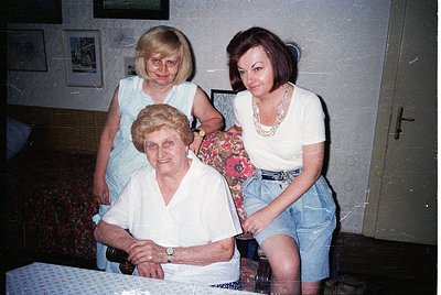 Three generations of women pose indoors, likely Eastern Europe, late 20th century. The seated woman wears a vintage white blo...