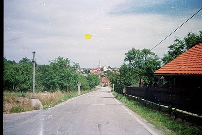 Vintage rural road scene with lush greenery, narrow asphalt path, and distant village rooftops. Sun flare highlights midday l...