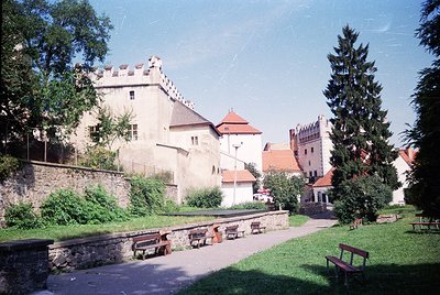 Medieval castle complex with stone towers, turrets, and fortified walls, surrounded by lush greenery. Prominent wooden benche...