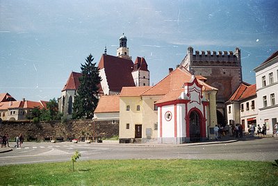 Historic European town square featuring a mix of medieval and Baroque architecture. Prominent red-brick tower with crenellati...
