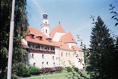 Historic castle with red-tiled gables and central tower, surrounded by lush greenery. Likely European architectural style, po...