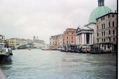 Venetian canal scene with historic architecture: domed church, arched bridges, and moored gondolas. Overcast sky enhances atm...