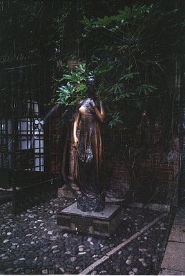 Bronze statue of draped female figure in contemplative pose, seated on stone pedestal in shaded courtyard. Iron bars and gree...