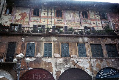 Faded Renaissance-style building façade featuring frescoed upper balcony with mythological figures, aged plaster, and arched ...