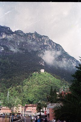 Historic castle perched atop lush green hillside, framed by dramatic alpine peaks. Foreground features Mediterranean-style bu...
