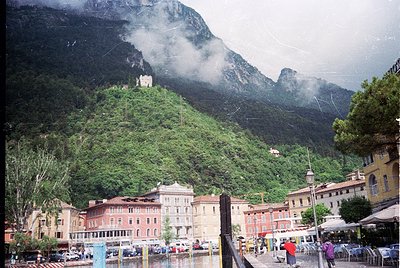 Alpine lakeside village nestled between dramatic rock formations and lush greenery. Colorful, multi-story buildings with Euro...