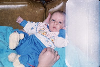 Vintage-style photo of an infant in a hospital bassinet, wearing a striped onesie and holding a white teddy bear. Soft focus ...