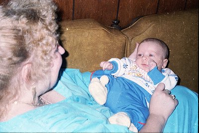 A tender moment captured: an elderly woman cradles a baby in a vintage-style blue onesie with white polka dots, seated on a w...