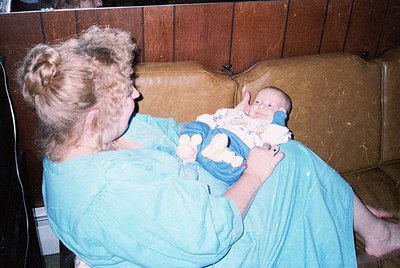Vintage candid of an adult cradling a swaddled infant in a worn leather armchair, likely mid-20th century. The woman’s hair i...