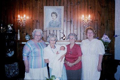 Four women pose indoors beside a framed portrait of a woman in a formal setting. The central woman holds a baby, while others...