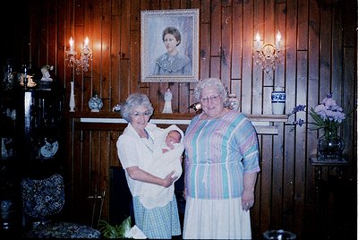 Two elderly women pose indoors with a newborn in a vintage-style home, likely 1970s–1980s. Wood-paneled walls, ornate wall sc...